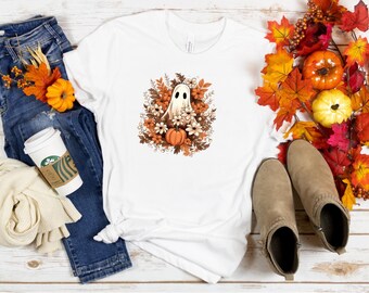 May include: White t-shirt featuring a ghost design surrounded by flowers and pumpkins. The design is in shades of brown, orange, and white. The shirt is styled with blue jeans, fall foliage, and tan boots.