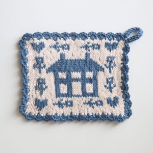 Op de afbeelding: Een blauwe en witte gebreide ovenwant met een huisontwerp. Het huis heeft een wit interieur en blauwe ramen en dak. De ovenwant heeft een blauwe rand met een gegolfde rand en een lus om op te hangen.