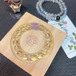 May include: Two bracelets made of natural stones. The first bracelet is made of yellow citrine stones and has a small purple stone charm. The second bracelet is made of white moonstone and has a black stone charm. Both bracelets are on a wooden display stand.