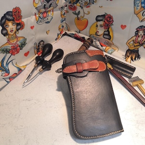 May include: A collection of leather crafting tools and accessories, including a dark gray leather case with a brown leather accent, awls, and a mallet. The background features a fabric with vintage tattoo-style illustrations.