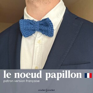 Könnte beinhalten: Eine blaue Häkel-Fliege, getragen über einem weißen Hemd und einem marineblauen Anzug. Der Text "le noeud papillon" und "patron version française" ist unten im Bild zu sehen.