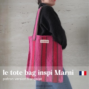Może przedstawiać: Różowo-brązowa torba typu tote wykonana na szydełku w paski z napisem "crochet crochet" na dole. Torbę trzyma osoba ubrana w czarny sweter i niebieskie dżinsy. Na dole obrazu widnieje napis "le tote bag inspi Marni".