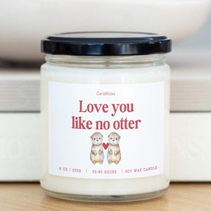 May include: A white soy wax candle with a black lid and a label that reads "CeraWicks Love you like no otter" with a graphic of two otters holding a red heart. The candle is 9 oz and has a burn time of 50-60 hours.