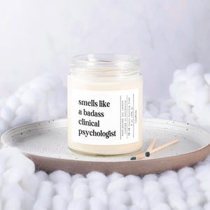 May include: A white candle in a glass jar with a label that reads "smells like a badass clinical psychologist".