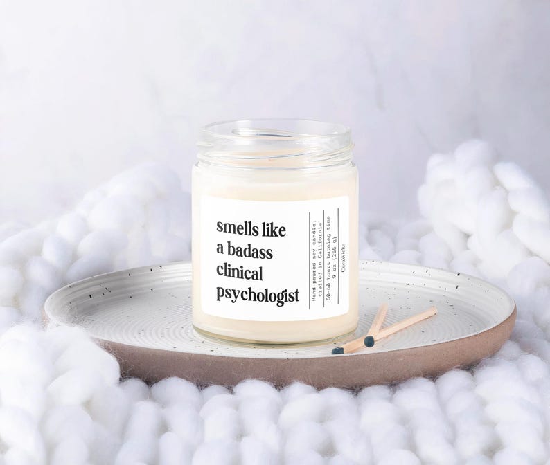 May include: A white candle in a glass jar with a label that reads "smells like a badass clinical psychologist".