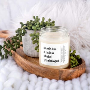 May include: A white soy candle with a black label that reads "smells like a badass clinical psychologist". The candle is on a wooden tray with a small green succulent plant in a gray pot.