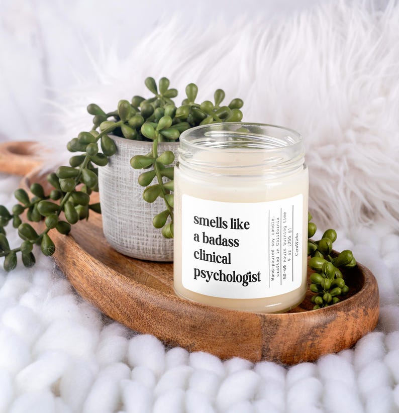 May include: A white soy candle with a black label that reads "smells like a badass clinical psychologist". The candle is on a wooden tray with a small green succulent plant in a gray pot.