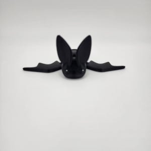 May include: A black bat-shaped decorative object with large ears and outstretched wings. The bat has a smooth, matte finish and is set against a plain white background. The object is likely a Halloween or gothic-themed decoration.