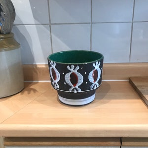 May include: A black ceramic plant pot with a green interior and a white and red pattern. The pot is on a light wooden surface. The pot's design is repeated around the circumference.