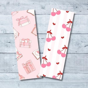May include: Two pink and white bookmark designs. One features a stack of books with pink ribbons and the other has a pattern of red cherries on a pink and white striped background.