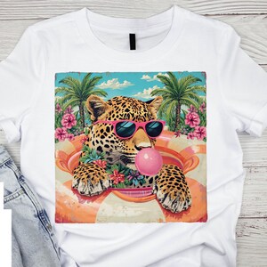 May include: White t-shirt featuring a leopard wearing pink sunglasses and blowing a bubblegum bubble. The leopard is in an orange and white inflatable ring, surrounded by flowers and palm trees. The design has a vintage, tropical vibe.
