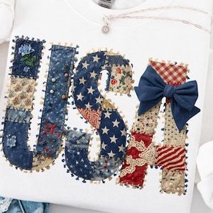 May include: White sweatshirt with the word "USA" appliqued in a patchwork design. The letters feature various patriotic patterns, including stars, stripes, and floral prints, with a blue bow on the "A". A silver necklace is visible.