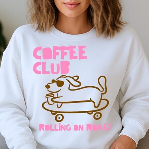 May include: White sweatshirt with the text "COFFEE CLUB" in pink. A brown cartoon dog wearing sunglasses and holding a coffee cup is on a skateboard. The text "ROLLING ON ROAST" is below the dog in pink.