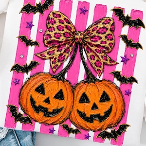 Op de afbeelding: Een wit sweatshirt met een grafische print van twee oranje pompoenen met een grote strik in luipaardprint. Het ontwerp bevat verticale felroze strepen, zwarte silhouetten van vleermuizen en kleine paarse sterren en doorzichtige steentjes.