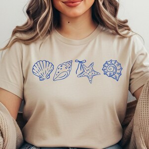 Puede incluir: Una camiseta beige con un dise&ntilde;o de conchas marinas azules. El dise&ntilde;o incluye una concha de vieira, una concha de caracola, una estrella de mar y una concha en espiral. La camiseta est&aacute; hecha de un material suave.