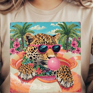 May include: A beige t-shirt featuring a colorful graphic of a leopard wearing pink sunglasses and blowing a bubblegum bubble. The leopard is in a life preserver with a tropical background of palm trees and flowers.