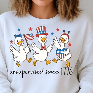 K&ouml;nnte beinhalten: Wei&szlig;es Sweatshirt mit drei Comic-G&auml;nsen, die den Unabh&auml;ngigkeitstag feiern. Die G&auml;nse sind mit patriotischen Accessoires wie einer Schleife, einem Zylinder und einem Herz geschm&uuml;ckt. Der Text "unsupervised since 1776" ist darunter gedruckt.