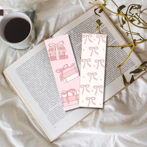 May include: Two pink bookmarks with a stack of books and pink bows on one and a repeating pattern of pink bows on the other. The bookmarks are on top of a book.