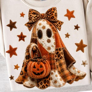 May include: A cream-colored sweatshirt featuring a large, central applique of a ghost wearing a leopard print bow. The ghost is constructed from various fabric patches in orange, brown, and cream, holding a small orange jack-o'-lantern bucket.
