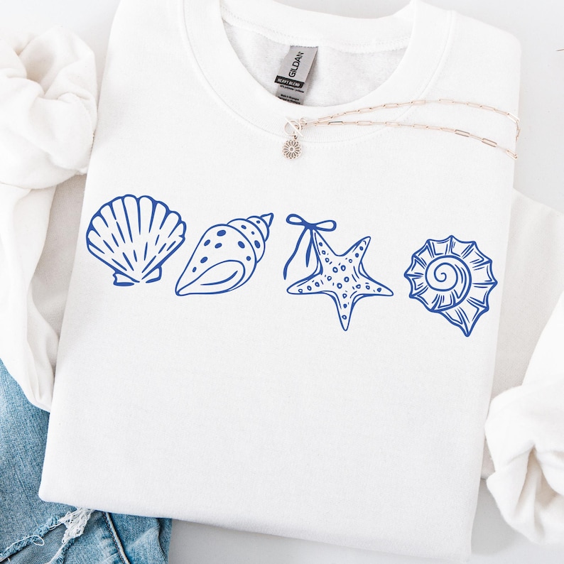 Puede incluir: Sudadera blanca con un dise&ntilde;o de conchas marinas azules. El dise&ntilde;o presenta cuatro ilustraciones diferentes de conchas marinas: una concha de vieira, una caracola, una estrella de mar y una concha en espiral. Un collar plateado con un peque&ntilde;o colgante.