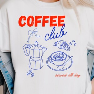 May include: White sweatshirt with a red and blue graphic. The graphic says "COFFEE club" and features a coffee pot, croissant, coffee beans, and a cup of coffee. The text "served all day" is also included.