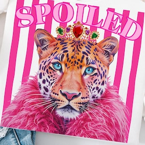May include: White t-shirt featuring a leopard illustration with blue eyes, wearing a jeweled crown and pink fur. The word "SPOILED" is printed in pink above the leopard, set against a background of pink stripes.