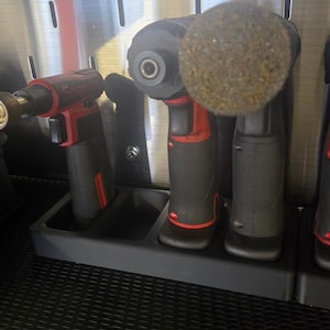 May include: Three power tools are stored in a black tool holder. The tools are a red and black cordless drill, a black cordless drill with a brown sanding pad, and a black and red torch. The tools are stored in a black tool holder with a black rubber mat underneath.