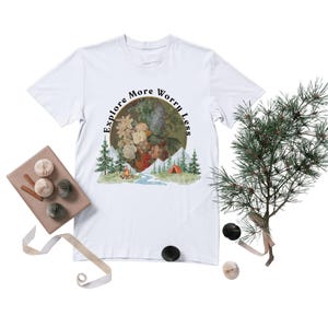 Peut inclure: T-shirt blanc avec un motif circulaire représentant des fleurs, des montagnes, une rivière et une tente. Le texte "Explore More Worry Less" est disposé en arc de cercle autour du motif floral. Éléments décoratifs : branche de pin, boîte cadeau et ornements en papier.