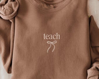 May include: A brown sweatshirt with the word "teach" in white script and a bow graphic. The long sleeves are rolled up, and the sweatshirt is displayed on a cream-colored surface. A hat and greenery are in the background.
