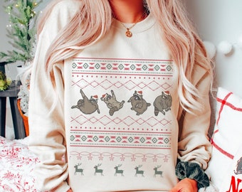 May include: Beige sweatshirt featuring a festive design with dancing raccoons wearing Santa hats. The design includes red and green geometric patterns and reindeer silhouettes. A cozy and fun holiday-themed apparel item.