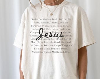 May include: Cream-colored t-shirt featuring the word "Jesus" in a stylized script. The shirt also displays a list of related words and phrases in a black font. The model is wearing black shorts and gold jewelry.