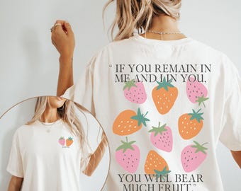 May include: Cream-colored t-shirt with a strawberry design. The back of the shirt features a cluster of pink and orange strawberries with green leaves, and the text "IF YOU REMAIN IN ME AND I IN YOU, YOU WILL BEAR MUCH FRUIT."