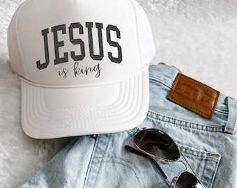 May include: White trucker hat with the text "JESUS is king" in black lettering. The hat is paired with light blue denim shorts and aviator sunglasses.