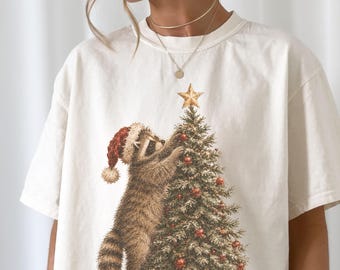 Op de afbeelding: Een off-white t-shirt met een grafische print van een wasbeer met een rood-witte kerstmuts die een gouden ster op de top van een versierde kerstboom met rode ornamenten plaatst. Het shirt heeft korte mouwen en een ronde halslijn.