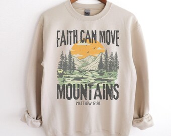 May include: Beige sweatshirt with a vintage-style graphic of a mountain landscape and the text "Faith can move mountains" with the Bible verse "Matthew 17:20" underneath.