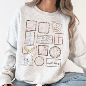 May include: Light gray sweatshirt with a collection of postage stamp-style illustrations in muted colors. The stamps depict various religious symbols and figures. The text "CHRIST IS KING" is printed at the bottom of the design.