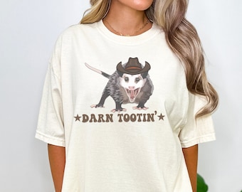 May include: A beige t-shirt with a graphic of a possum wearing a cowboy hat. The possum is looking at the viewer with its mouth open. The text "DARN TOOTIN'" is printed below the possum.