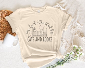 May include: A soft cream colored t-shirt with a black graphic design that says "easily distracted by cats and books". The graphic features a bookshelf with cats and plants.