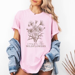 May include: A pink t-shirt with a brown line drawing of a bouquet of wildflowers and the text "consider the wildflowers".