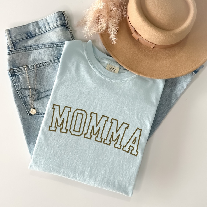 May include: A light blue t-shirt with the word "MOMMA" printed in brown block letters. The shirt is folded and laid on top of blue jeans and a brown hat.