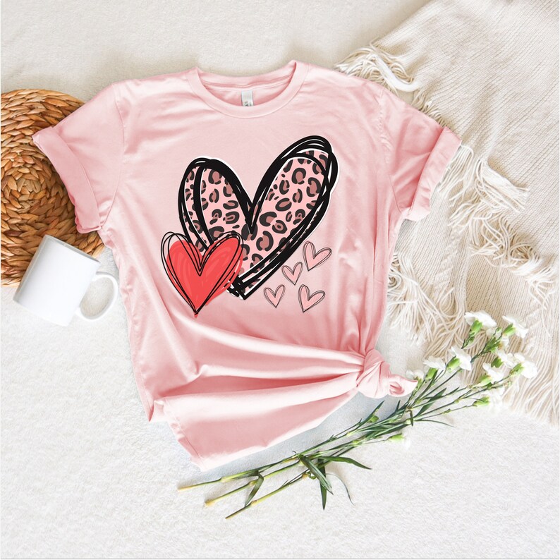 May include: Pink t-shirt with a black and white outline of two hearts. The larger heart has a leopard print pattern. A smaller red heart is inside the larger heart. There are four smaller pink hearts around the larger heart.