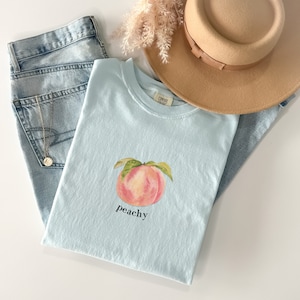 May include: A light blue t-shirt with a watercolor peach illustration and the text "peachy" printed on the front.