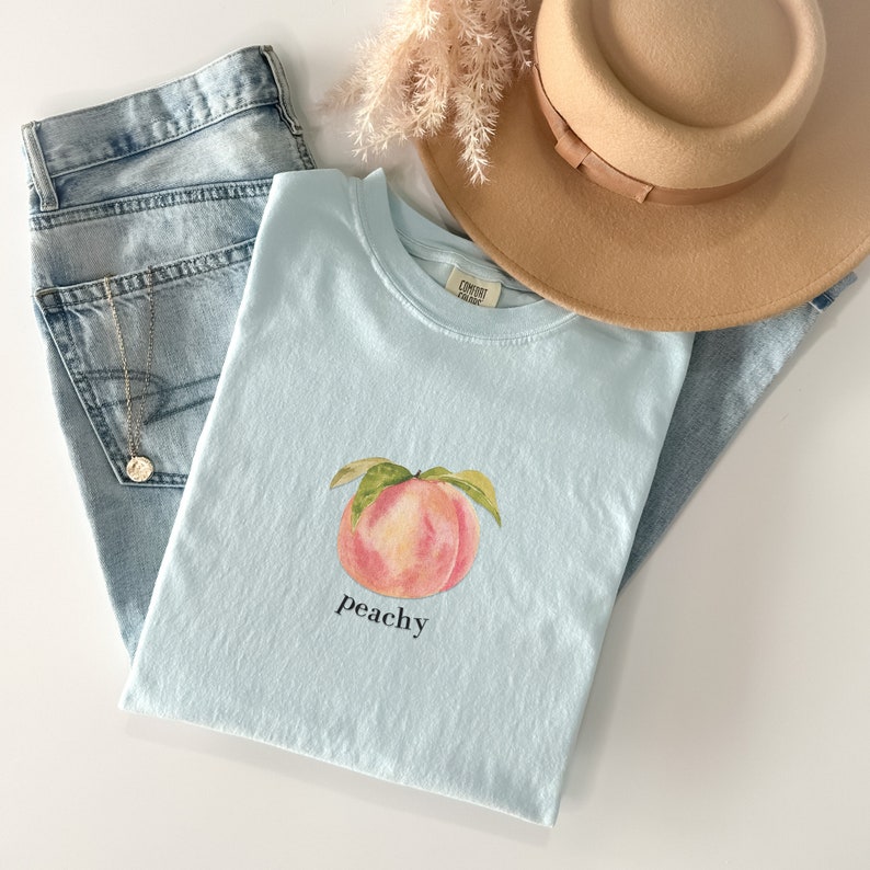 May include: A light blue t-shirt with a watercolor peach illustration and the text "peachy" printed on the front.