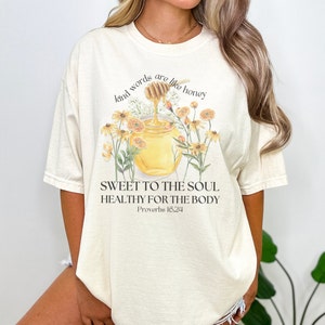 May include: A beige t-shirt with a graphic of a honey pot with a dripping honey dipper and flowers. The text "kind words are like honey" is above the graphic and the text "SWEET TO THE SOUL HEALTHY FOR THE BODY Proverbs 16:24" is below the graphic.