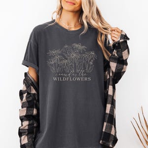 May include: A charcoal gray t-shirt with a white outline of wildflowers and the text "consider the wildflowers".