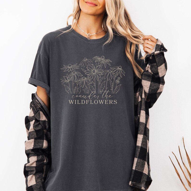 May include: A charcoal gray t-shirt with a white outline of wildflowers and the text "consider the wildflowers".