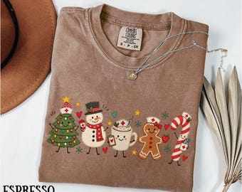 May include: An espresso-colored t-shirt featuring a graphic print of a Christmas tree, a snowman, a mug of cocoa, a gingerbread person, and a candy cane, all wearing nurse caps. The shirt has a Comfort Colors label at the inner neckline.