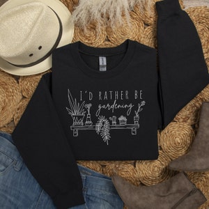 Peut inclure: Sweat-shirt noir avec un texte blanc qui dit "J'aimerais mieux jardiner" et un graphique de plantes en pot sur une &eacute;tag&egrave;re.