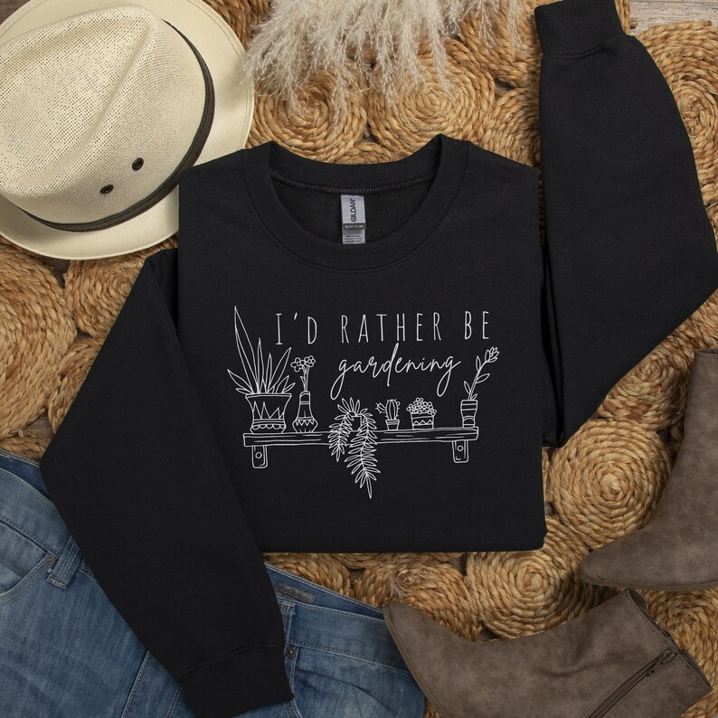 Peut inclure: Sweat-shirt noir avec un texte blanc qui dit "J'aimerais mieux jardiner" et un graphique de plantes en pot sur une &eacute;tag&egrave;re.