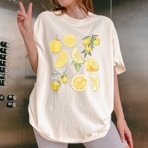 Pu&ograve; includere: Una maglietta color crema con una stampa grafica di limoni e fette di limone. I limoni sono gialli e le foglie sono verdi.